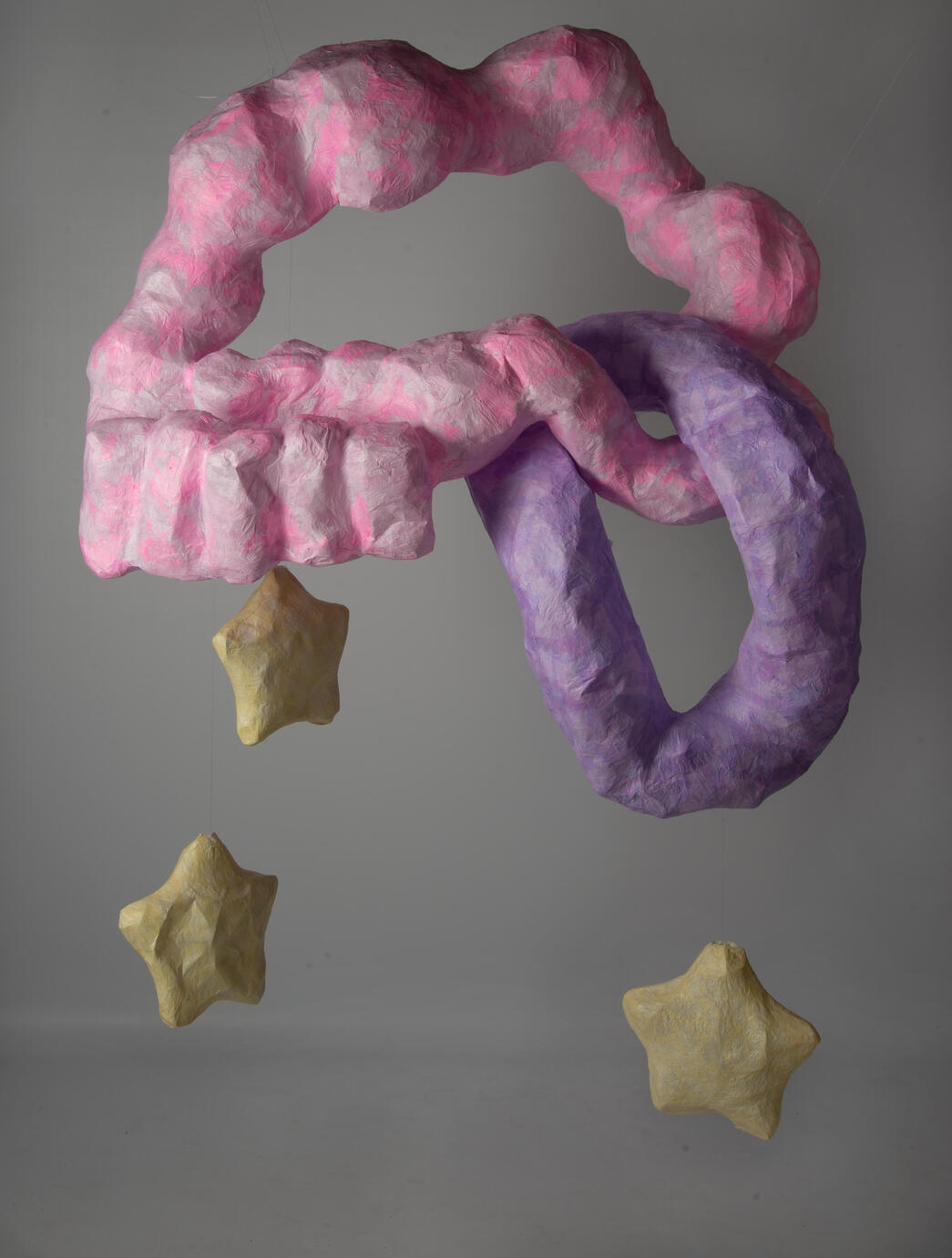 A hanging paper mache sculpture made up of a large pink amorphous looped shaped with a smaller purple part looping through it, Three yellow paper mache stars hang from the sculpture.