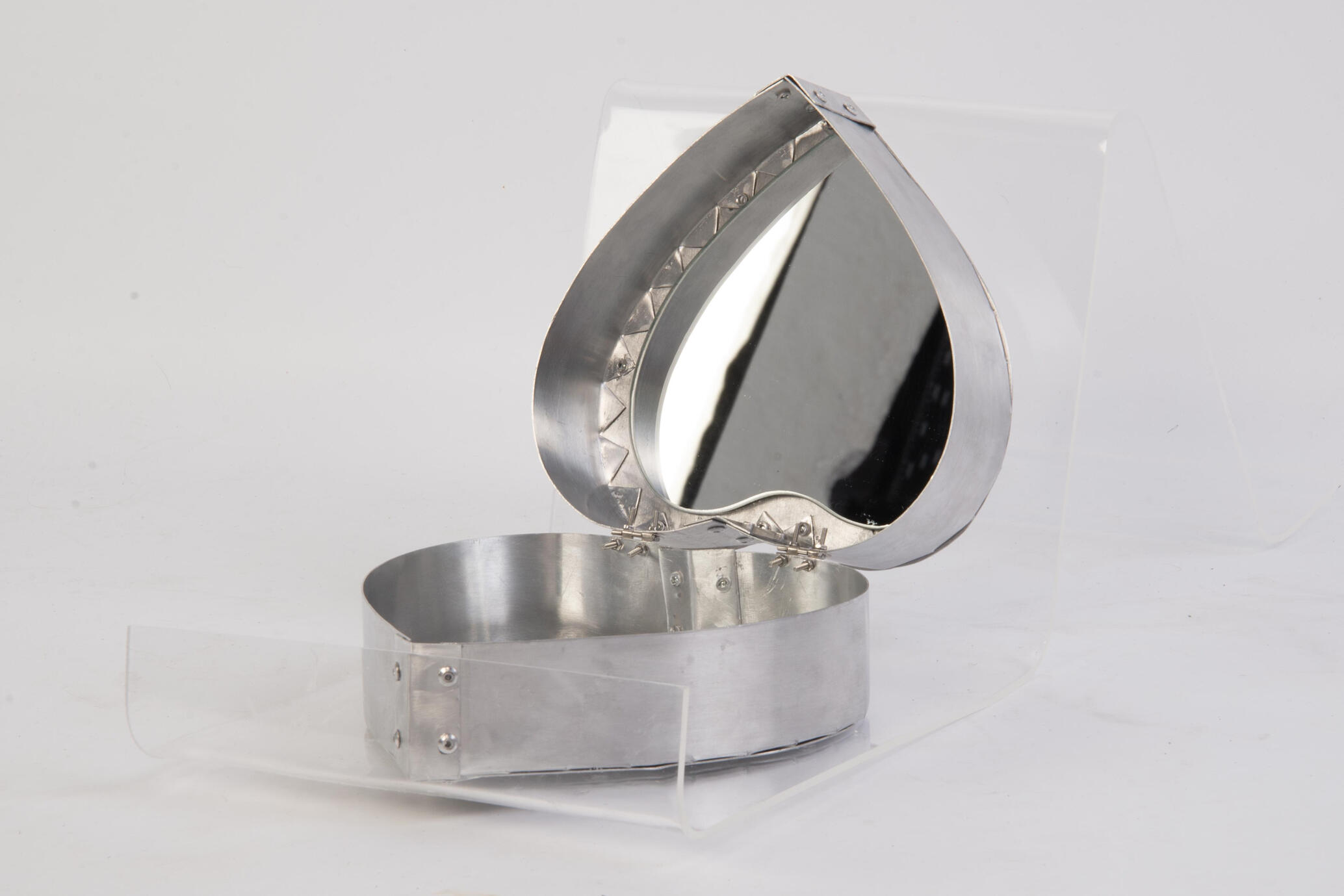 A heart shaped aluminium box with visible rivets and bolts, with pointed aluminium tabs surrounding a heart shaped mirror on the inside.