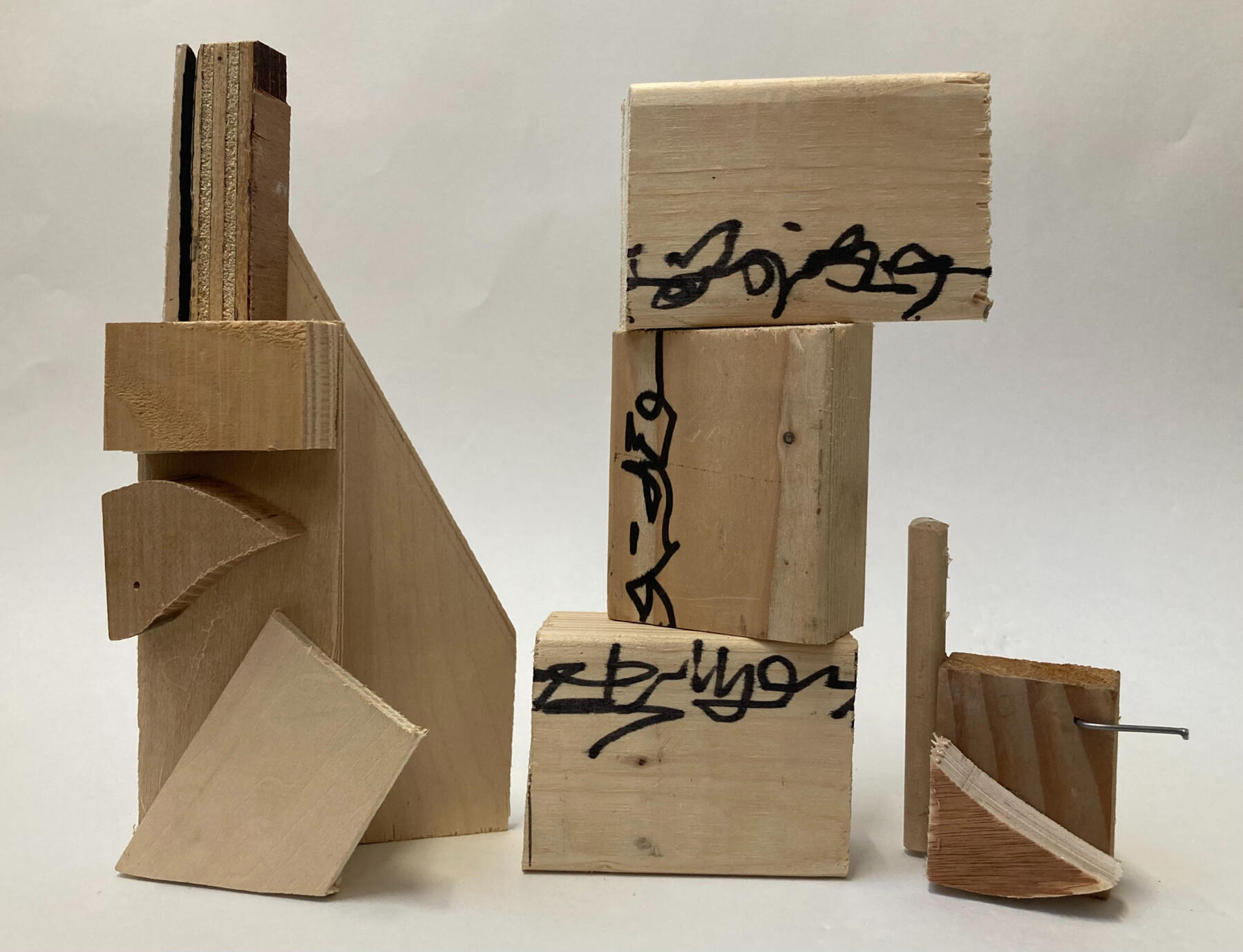 Three wooden block sculptures. The left is made up of glued together wooden pieces forming a triangle-like outline, the middle is 3 blocks stacked with scribbled markings across them, the third is smaller and is made up of 3 pieces of wood and a nail.