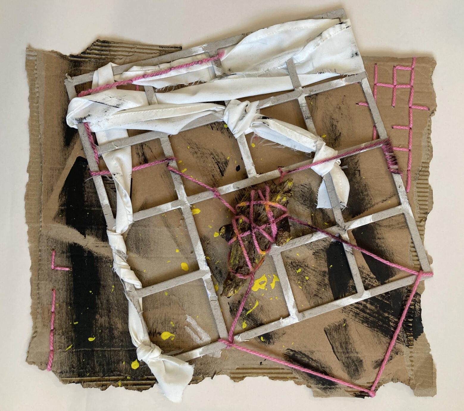 A cardboard rectangle with ripped edges, painted with abstract black marks and yellow paint splatters. There is a grid of thick cardboard attached with knotted white fabric. The piece is stitched into with pink yarn creating lines and squares.