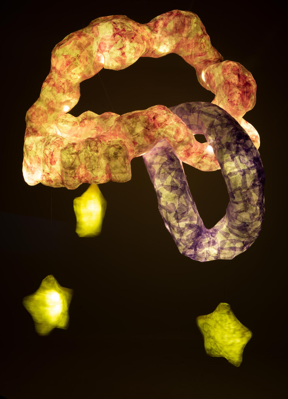 A hanging paper mache sculpture lit from within against a black background. It is made up of a large pink amorphous looped shaped with a smaller purple part looping through it, Three yellow paper mache stars hang from the sculpture.