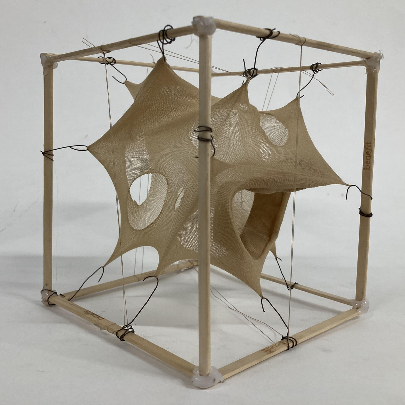A cube shaped frame of smooth, flat wooden sticks contains a holey, pale tan nylon wig cap that is being stretched to different points on the frame using wire hooks and brown thread.