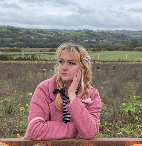 A head and shoulders photo of a blonde women in her 20s looking to the left, against a background of English fields and hills. Her hair is in two braids with bows and and she is wearing a pink coat.