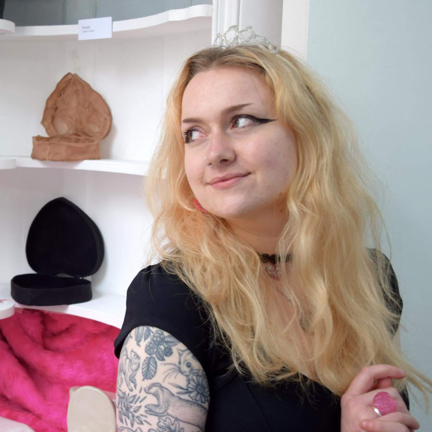 A head and shoulders photo of a blonde woman in her 20s looking to the left in front of her art.