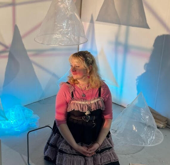 A blonde woman in her 20s looking to the left in front of her art.